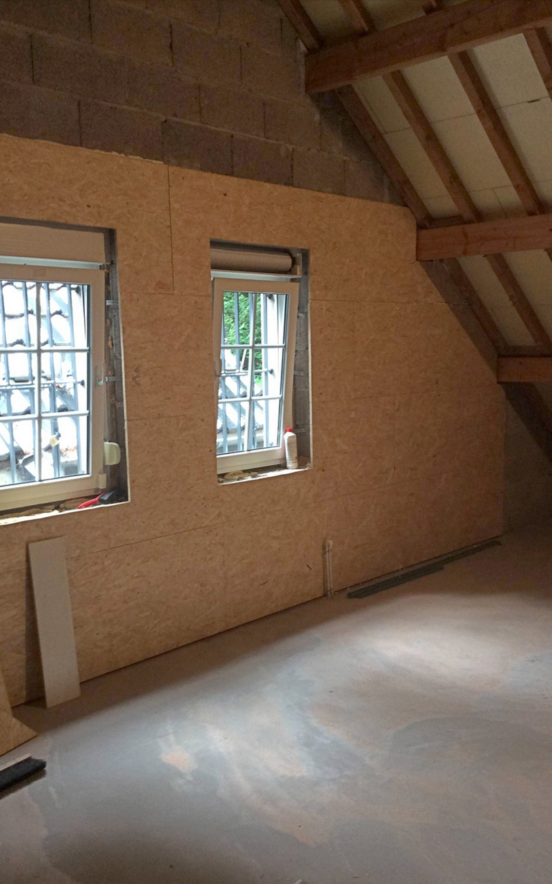 Innenausbau eines Dachgeschosses mit OSB-Platten verkleideten Wänden, zwei Fenstern mit Sprossen und sichtbaren Holzbalken der Dachkonstruktion. Der Raum ist hell und noch unbewohnt.